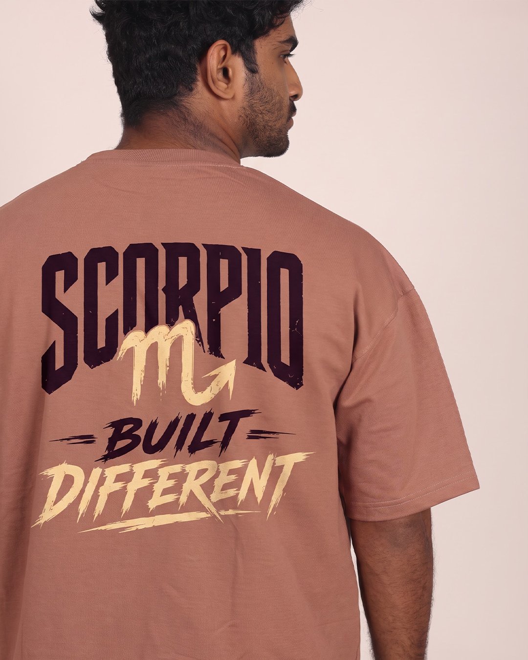 Man wearing Rhino Rave brown oversized T-shirt featuring a large “SCORPIO – BUILT DIFFERENT” graphic on the back.